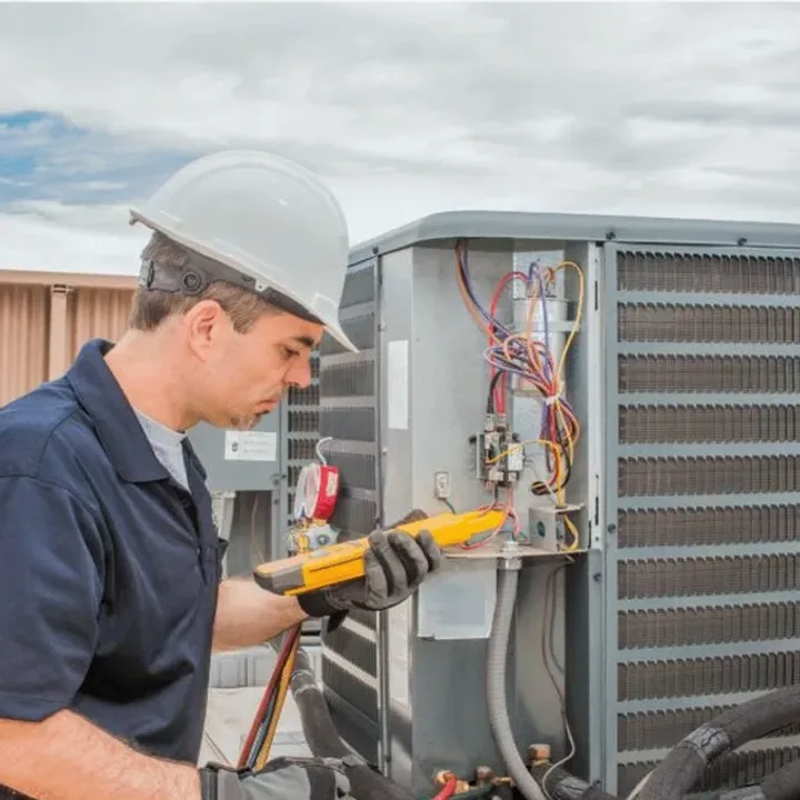 HVAC technician in a hard hat testing an outdoor air conditioner with a multimeter